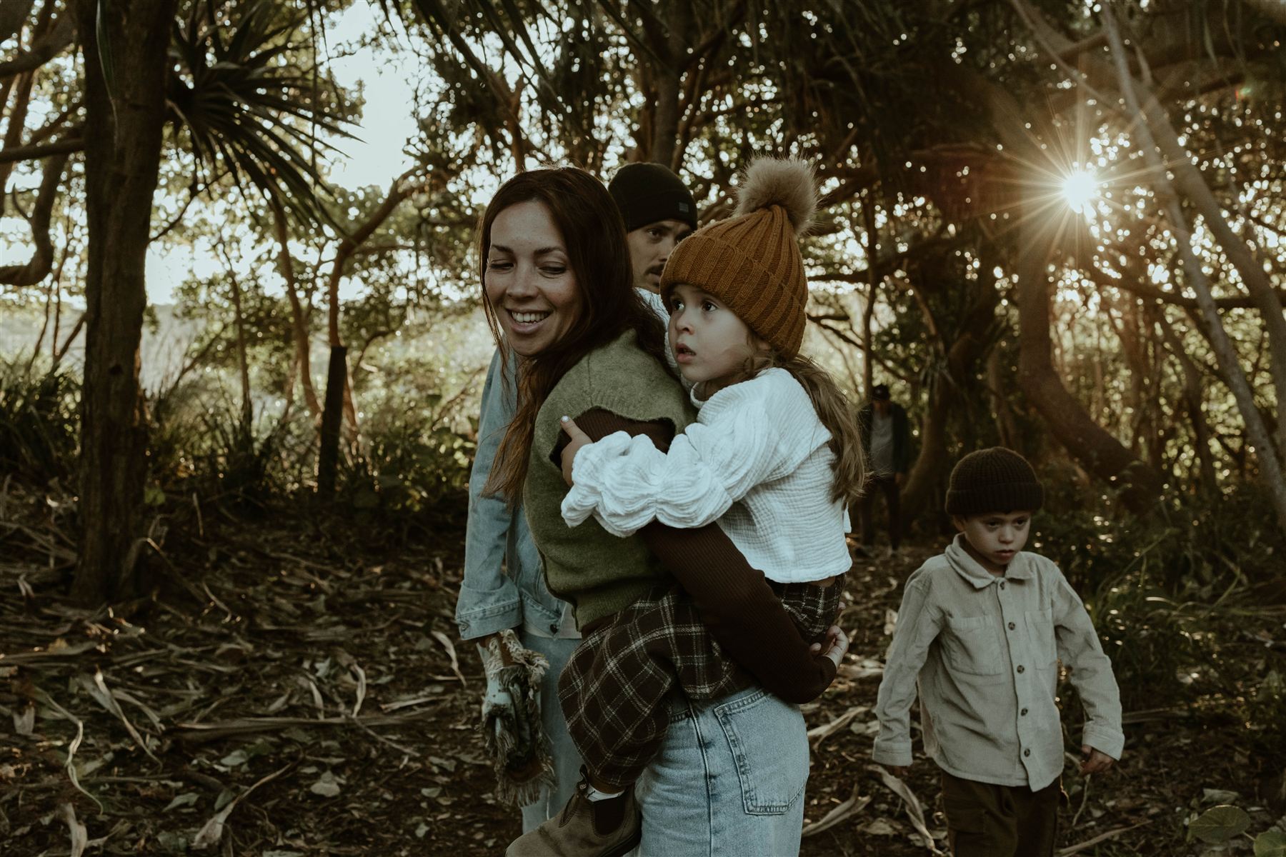 Family walking through coastal forest at golden hour, mother carrying her daughter