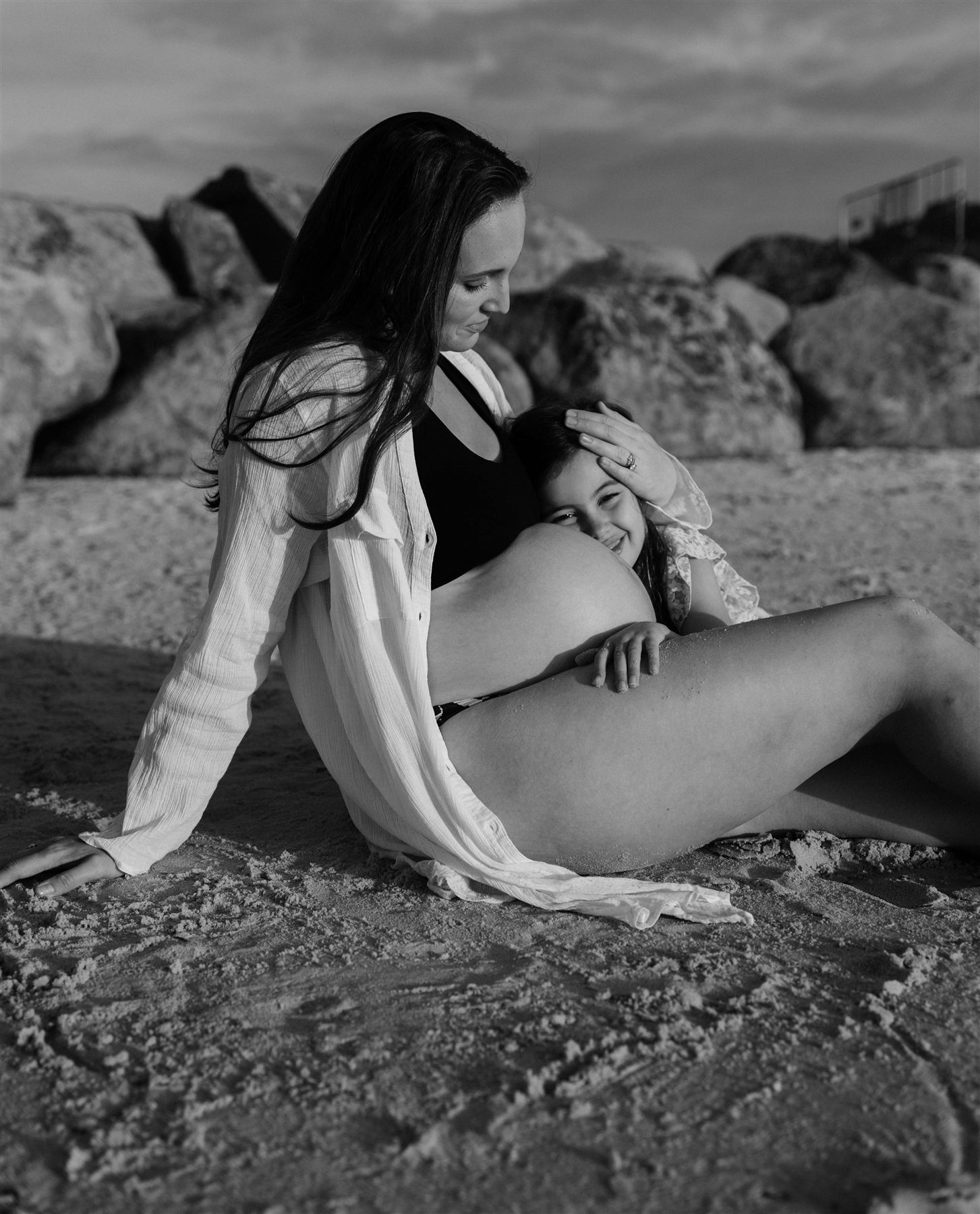 Black and white photograph of a daughter cuddling her mother's pregnant belly on the beach